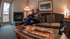 Un salon chaleureux avec une cheminée, un canapé et une table où sont disposés des en-cas et une bouteille de vin.