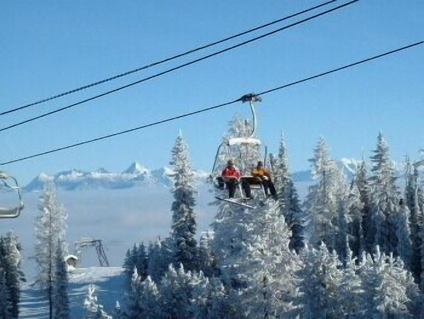 Un télésiège avec deux passagers, entouré d’arbres couverts de neige et avec des montagnes enneigées en arrière-plan.