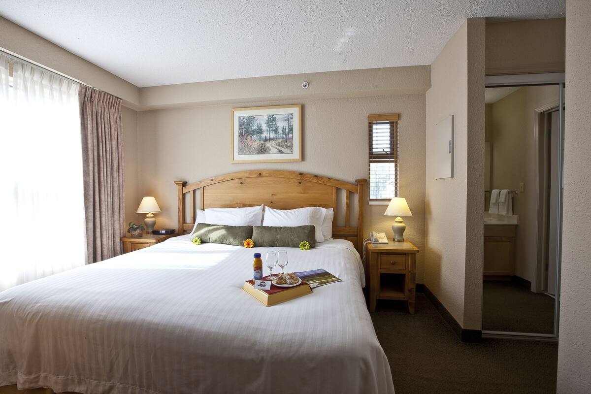 Habitación de hotel con una cama grande, dos mesitas de noche con lámparas, un cuadro enmarcado en la pared y una puerta que da a un baño.