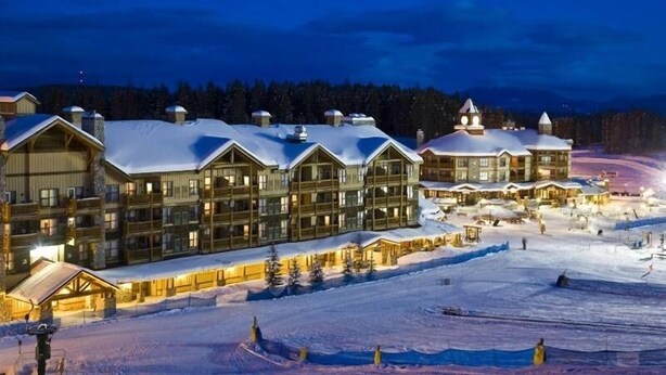 Un chalet composé de plusieurs bâtiments en bois, avec des toits enneigés, et une piste de ski au premier plan.