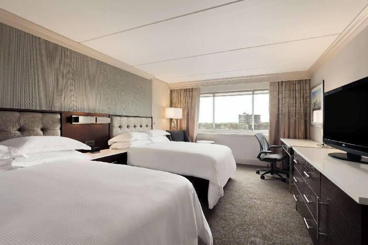 A hotel room with two beds, a desk with a TV, a chair, and a window with a city view.