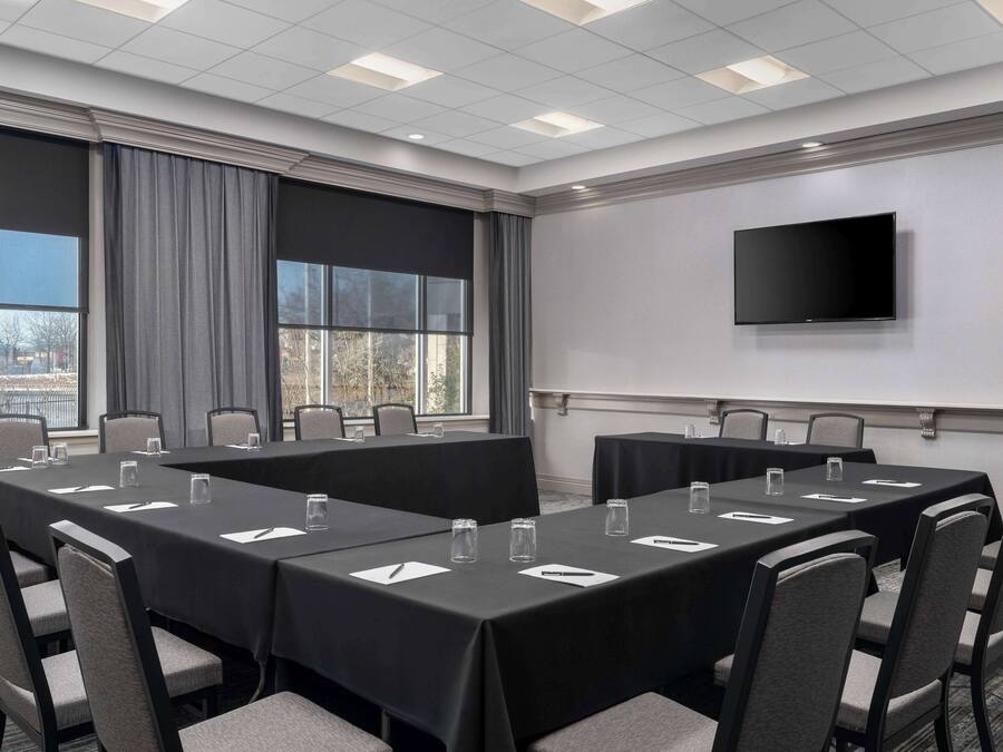 Uma sala de conferências com uma mesa grande, cadeiras, uma TV de tela plana e janelas com cortinas.