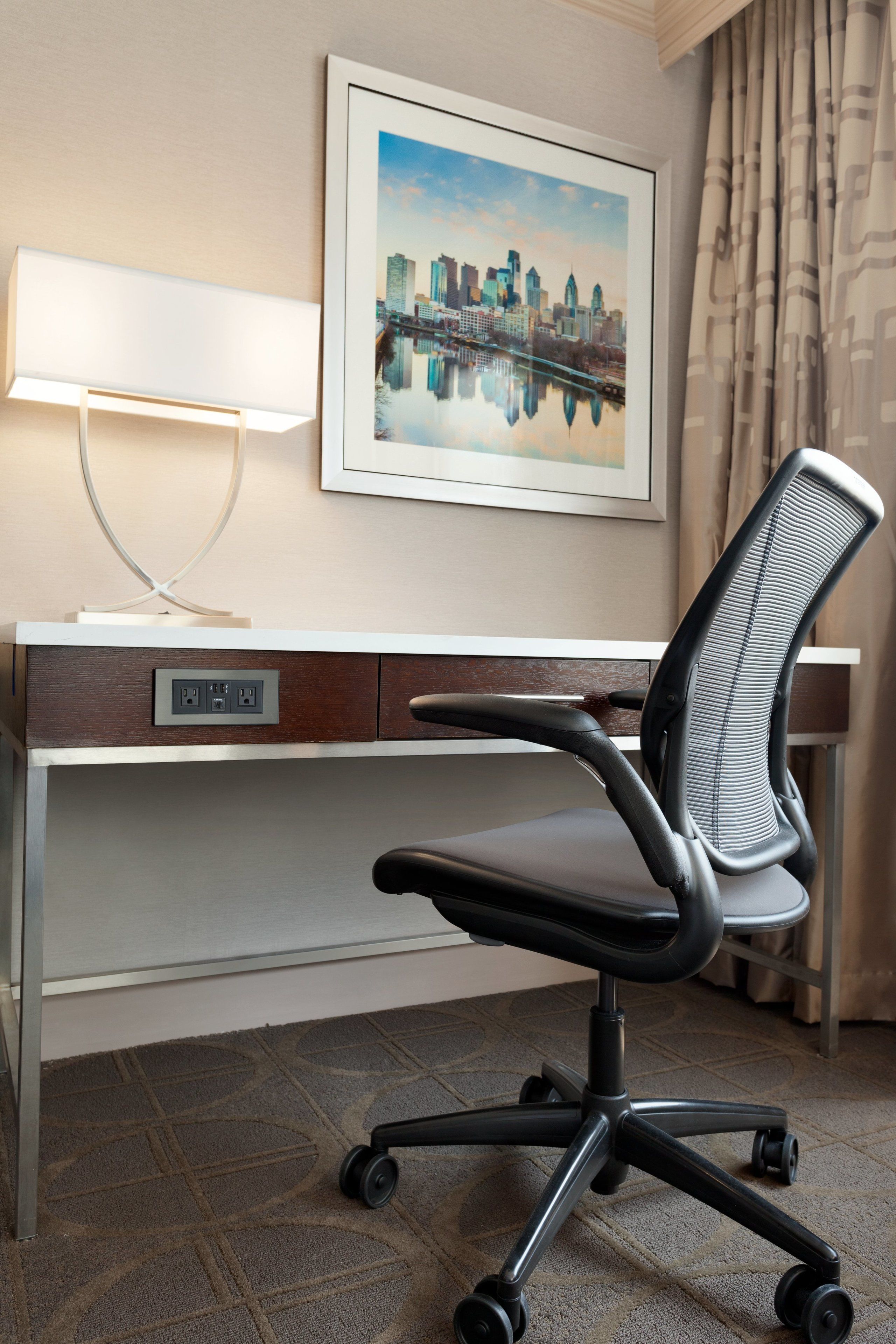 A hotel room with a desk, a chair, a lamp, and a framed cityscape picture on the wall.
