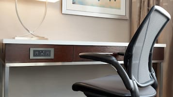 A hotel room with a desk, a chair, a lamp, and a framed cityscape picture on the wall.