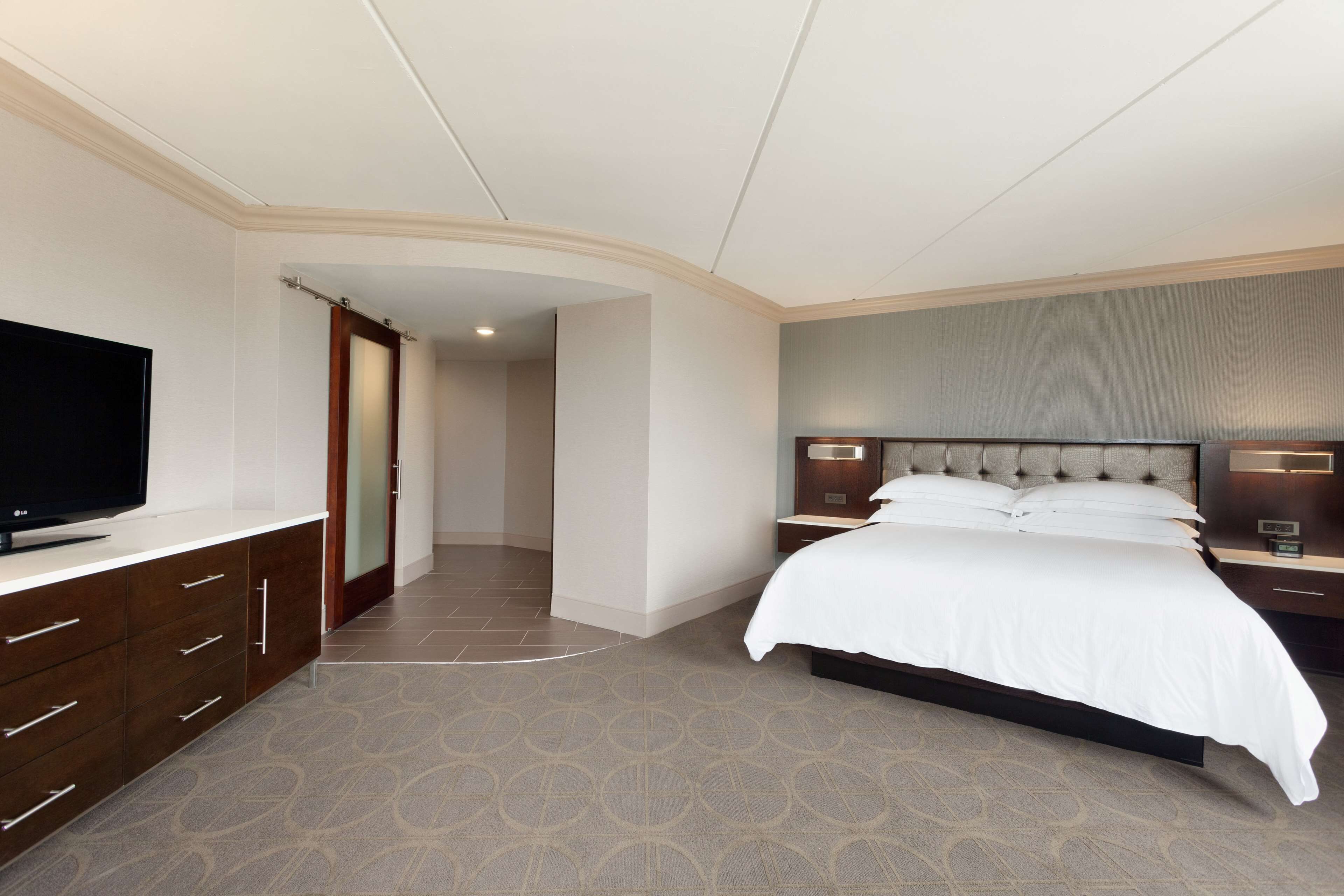 A modern hotel room with a large bed, a flat-screen TV, a wooden dresser, and a walk-in closet.
