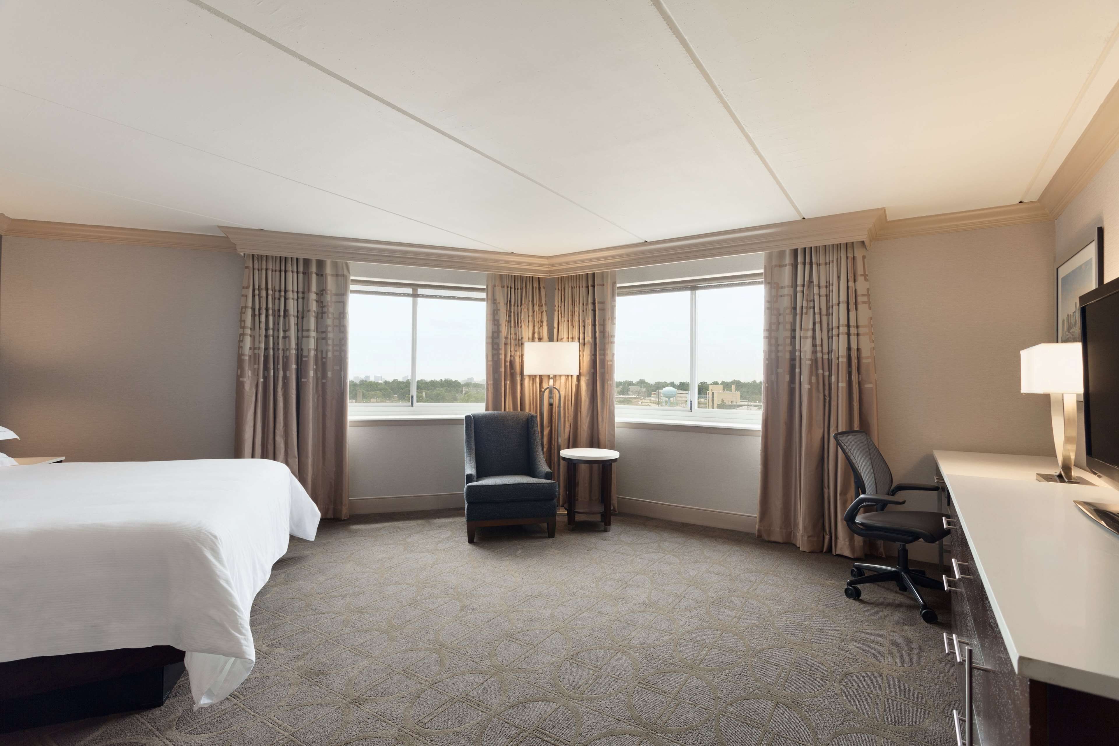 A hotel room with a bed, a desk, a chair, a lamp, and large windows.