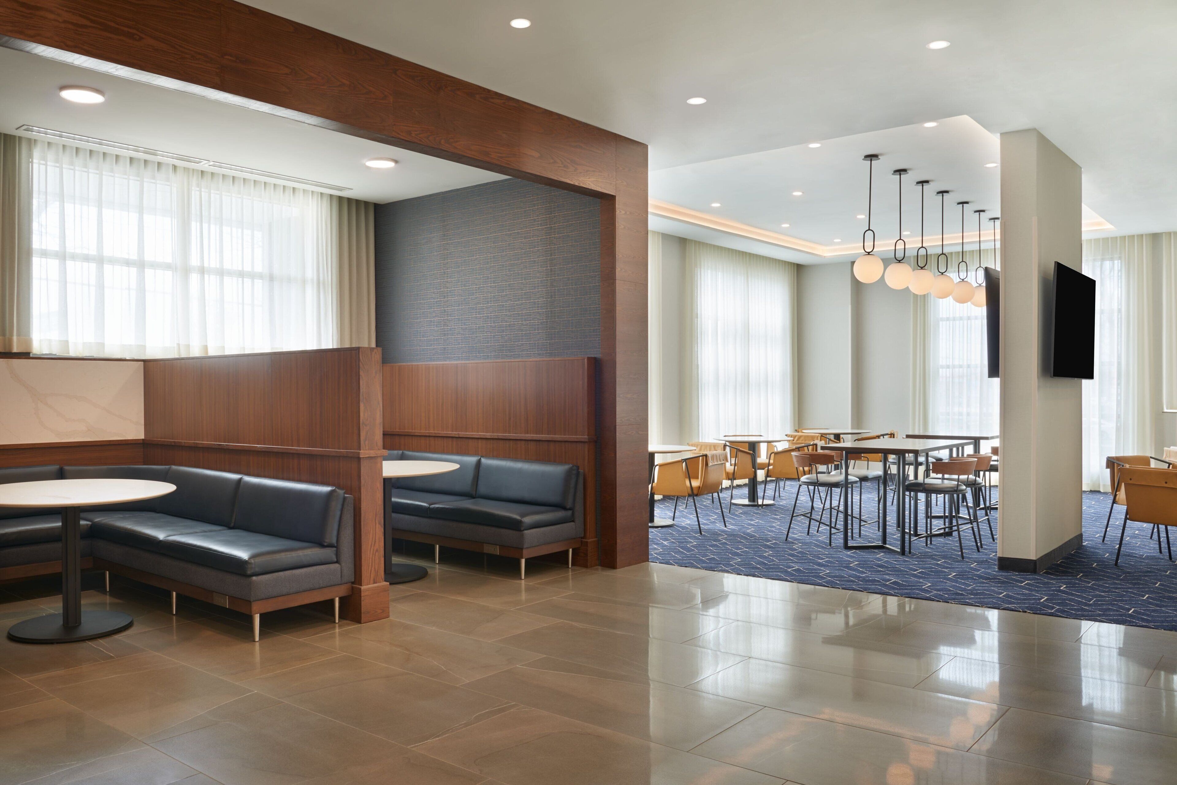 A modern hotel lobby with a seating area, a dining section, and a flat-screen TV mounted on the wall.