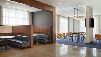 A modern hotel lobby with a seating area, a dining section, and a flat-screen TV mounted on the wall.