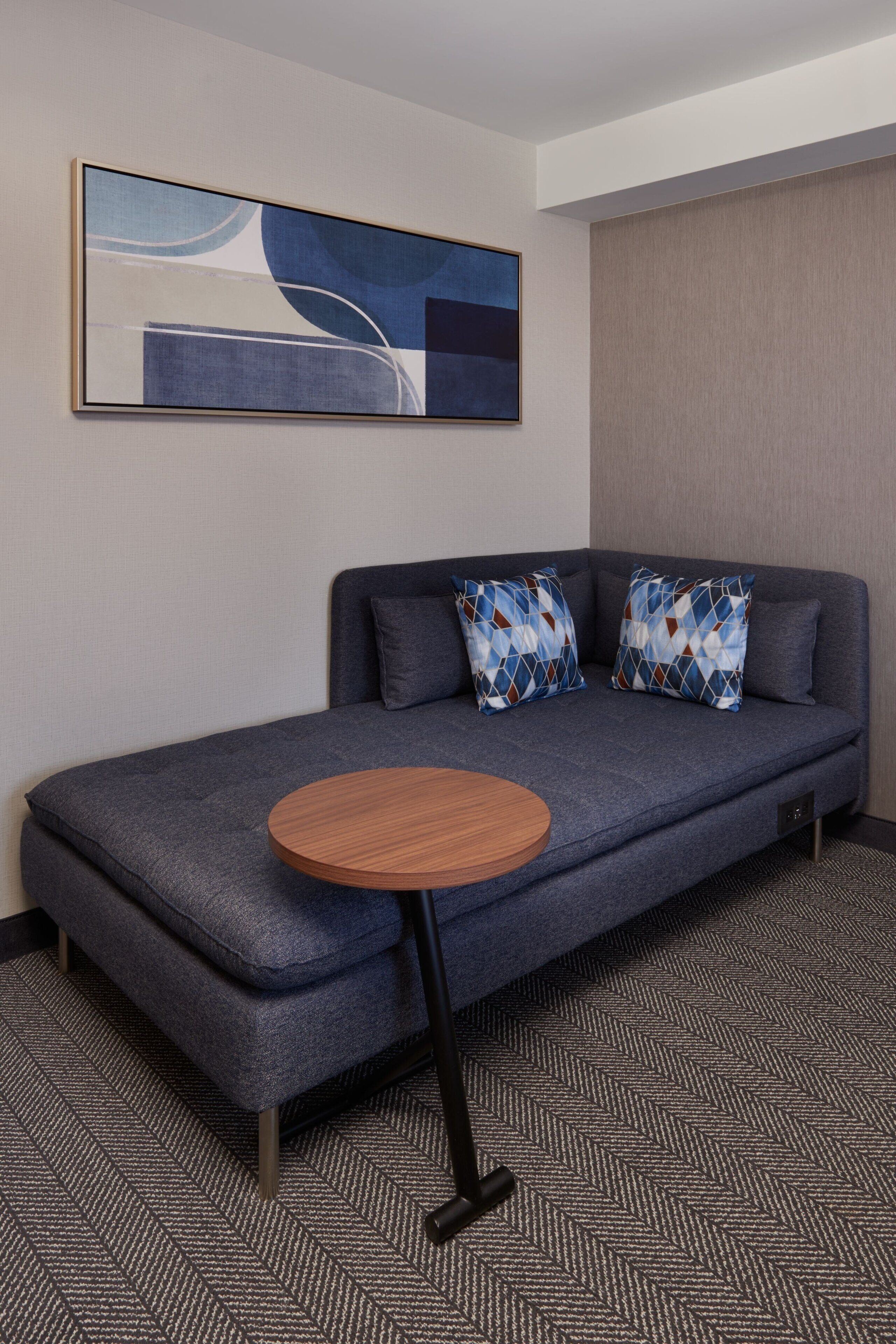 Une chambre d’hôtel moderne avec un canapé gris, deux coussins à motifs et une petite table d’appoint ronde en bois.