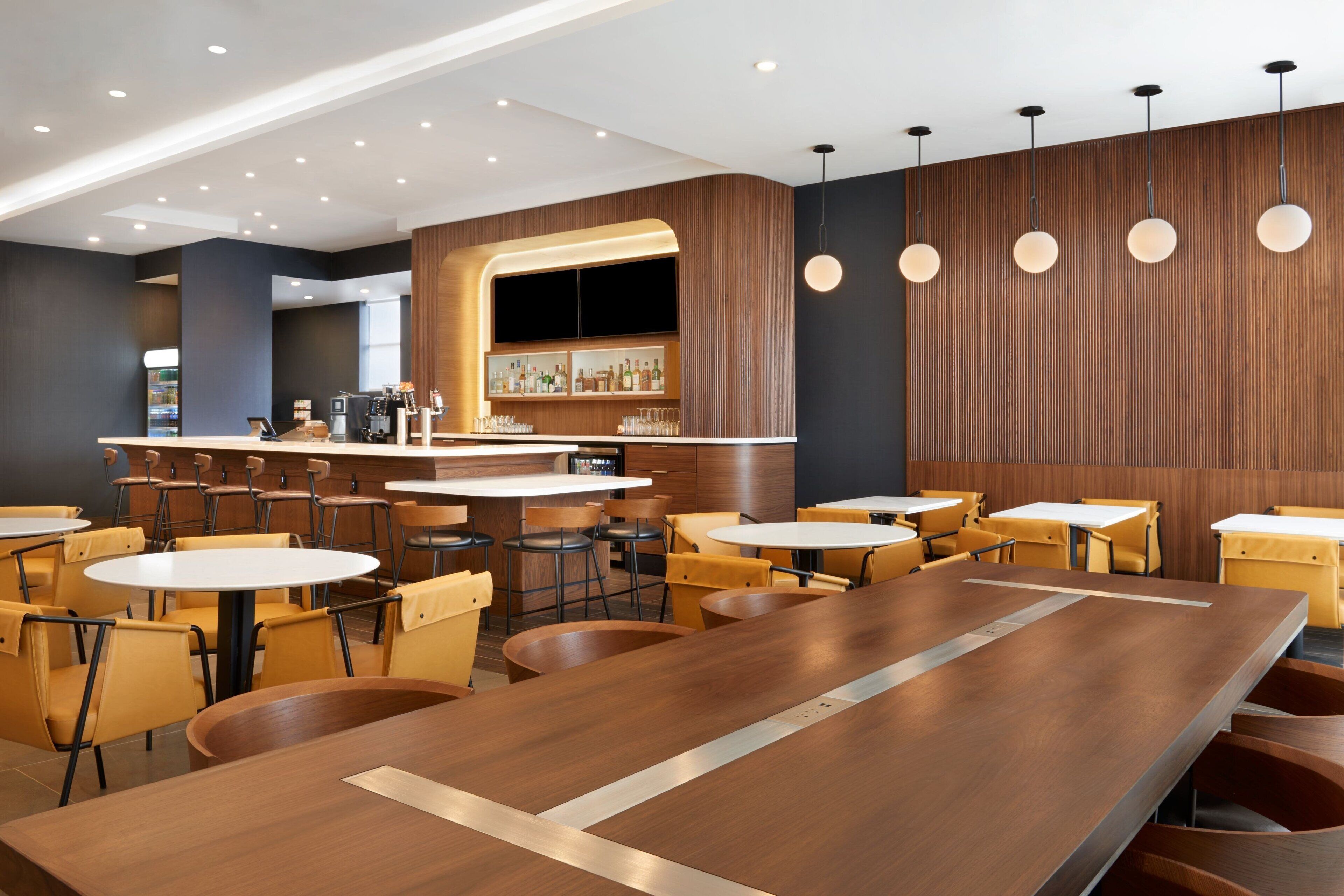 Um restaurante moderno com mesas e cadeiras de madeira, área de bar e iluminação pendente.