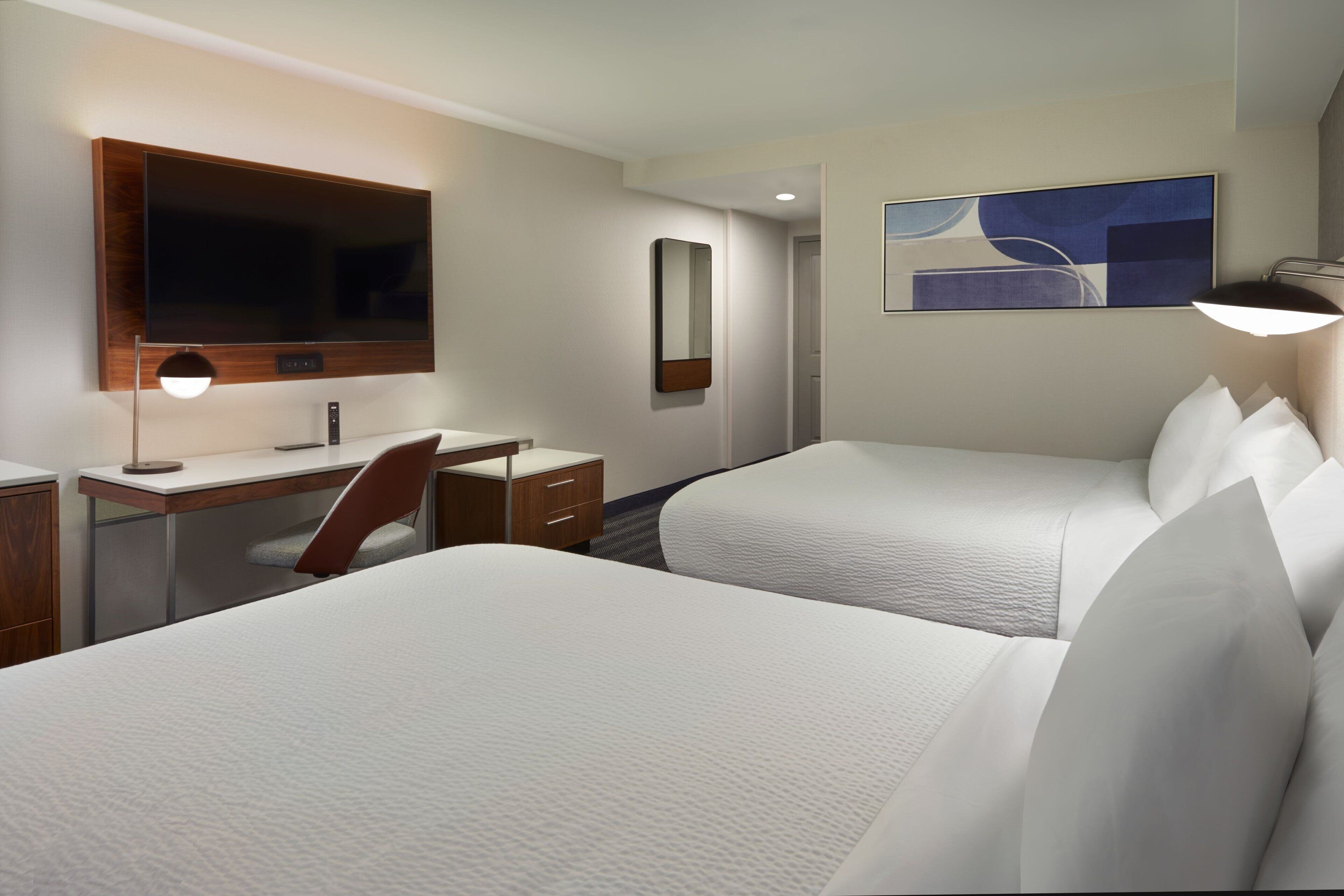 A hotel room with two beds, a desk, a chair, a TV, a mirror, and a painting on the wall.