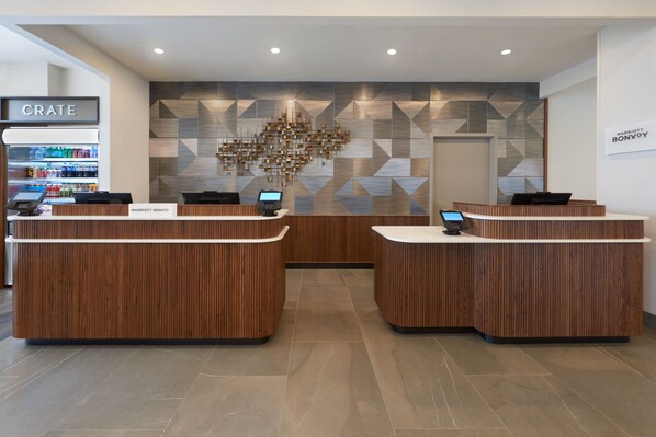 A hotel reception area with a modern design, featuring a central desk with a digital screen, two smaller desks, and a 'CRATE' refrigerator with beverages. - Courtyard by Marriott Kitchener (Kitchener)