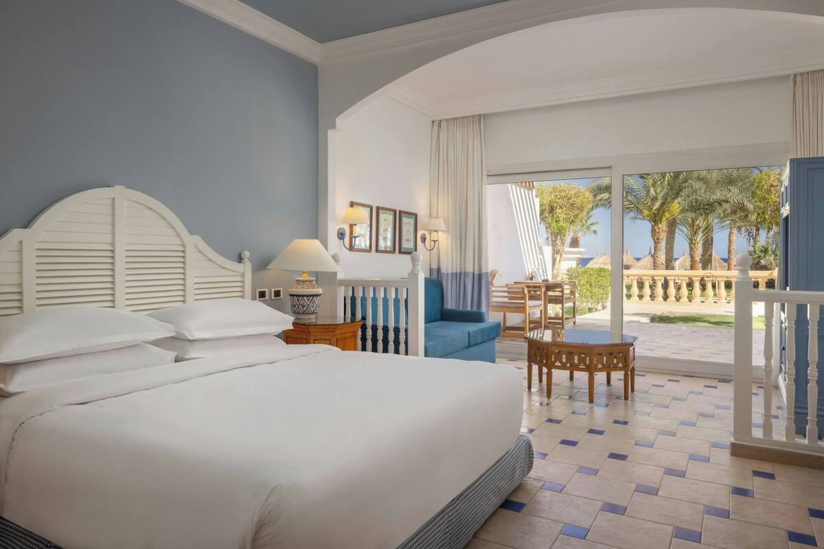 A hotel room with a large bed, a balcony with a table and chairs, and a view of palm trees.