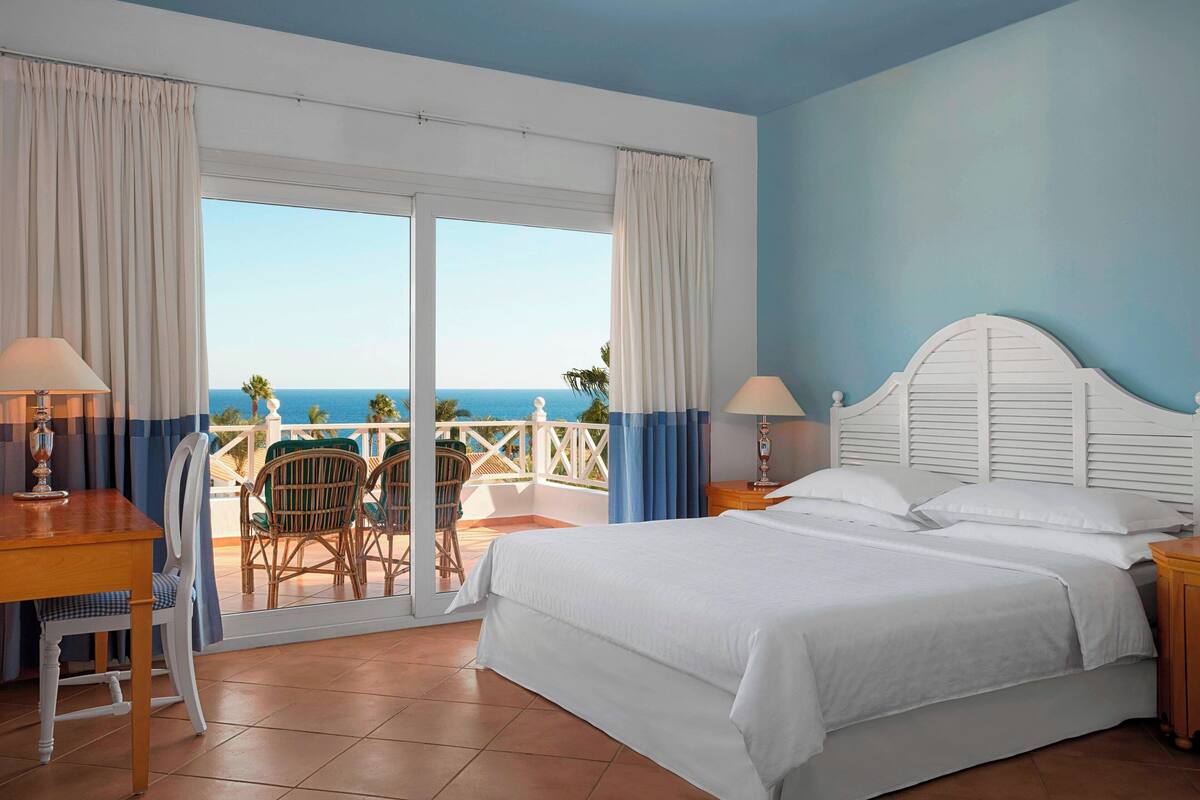 A bedroom with a large bed, a desk, and a chair. There is a balcony with a view of the ocean.