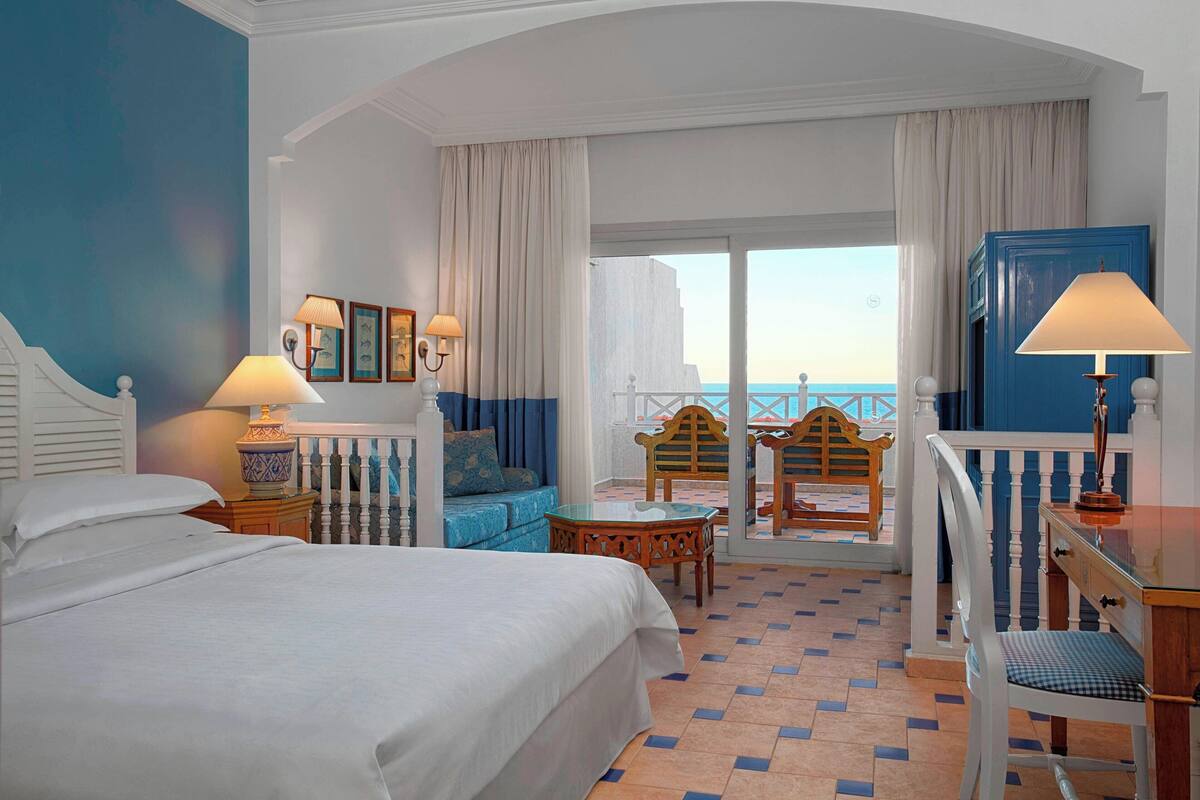 A hotel room with a bed, a desk, a chair, a sofa, and a balcony with a view of the sea.