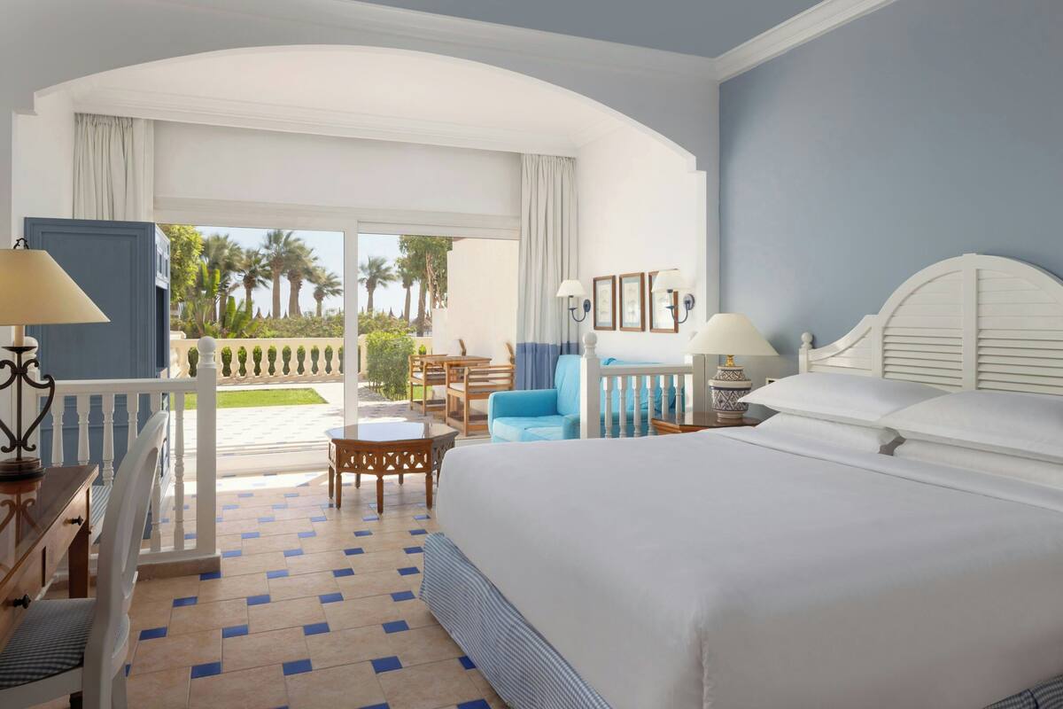 A hotel room with a bed, a desk, a chair, a small table, a lamp, a nightstand, and a view of the outdoors.