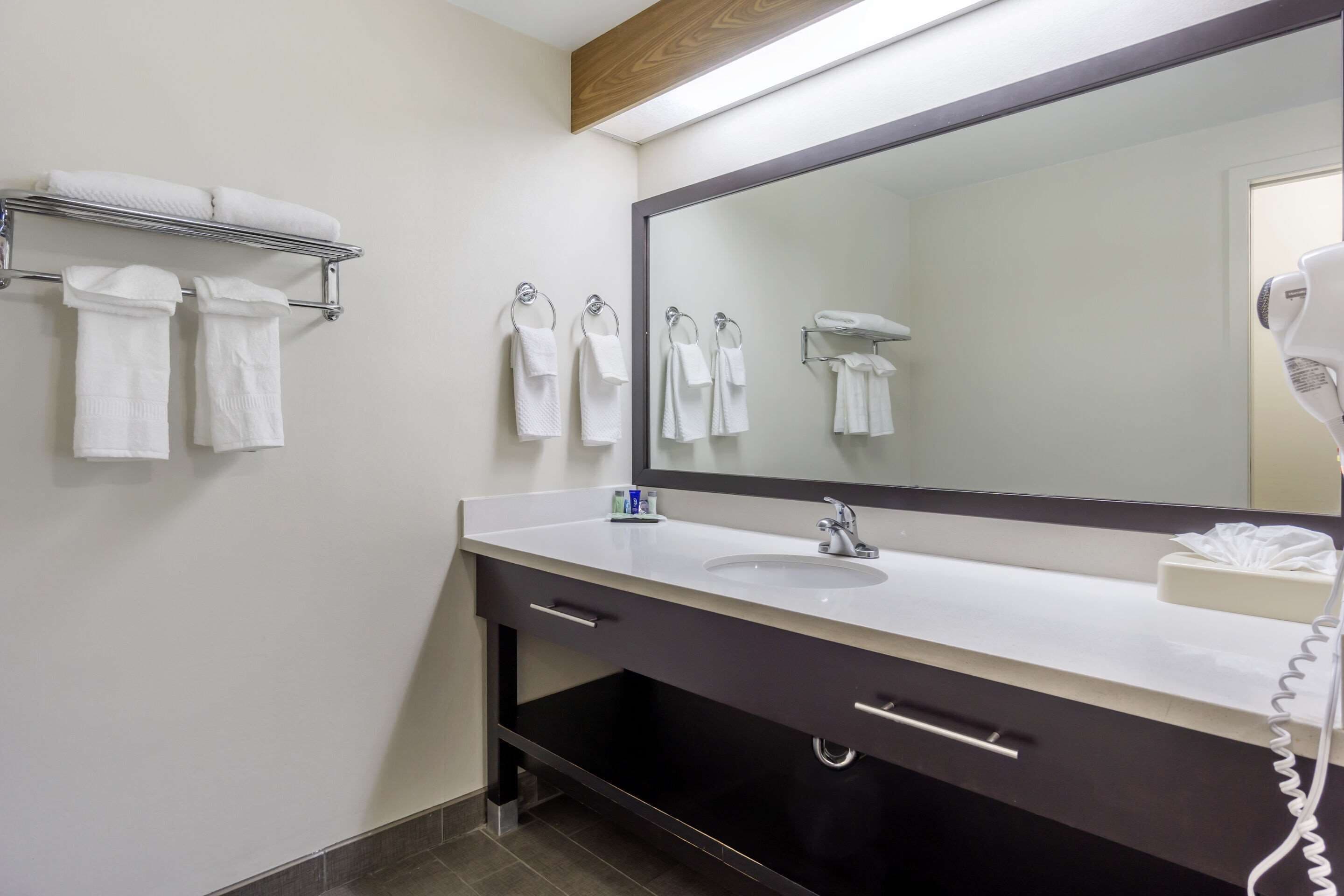 Une salle de bain moderne avec un grand miroir, un lavabo blanc, un sèche-cheveux et plusieurs serviettes rangées sur un porte-serviettes.