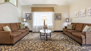 A hotel room with two brown sofas, a round coffee table with a vase of flowers, a large window with sheer curtains, and framed artwork on the walls. - Sleep Inn - SeaTac Airport (SeaTac)