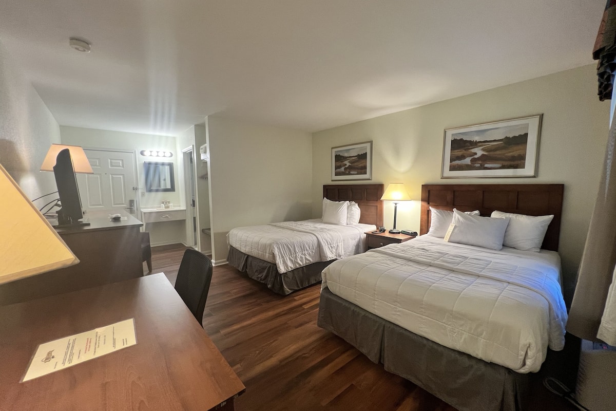 A hotel room with two beds, a desk, a chair, a lamp, and a painting on the wall.