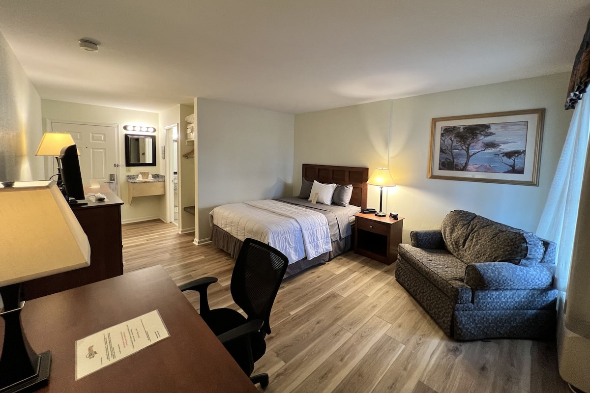 A hotel room with a bed, a desk with a lamp, a chair, a nightstand with a lamp, a painting on the wall, and a bathroom visible through an open door.