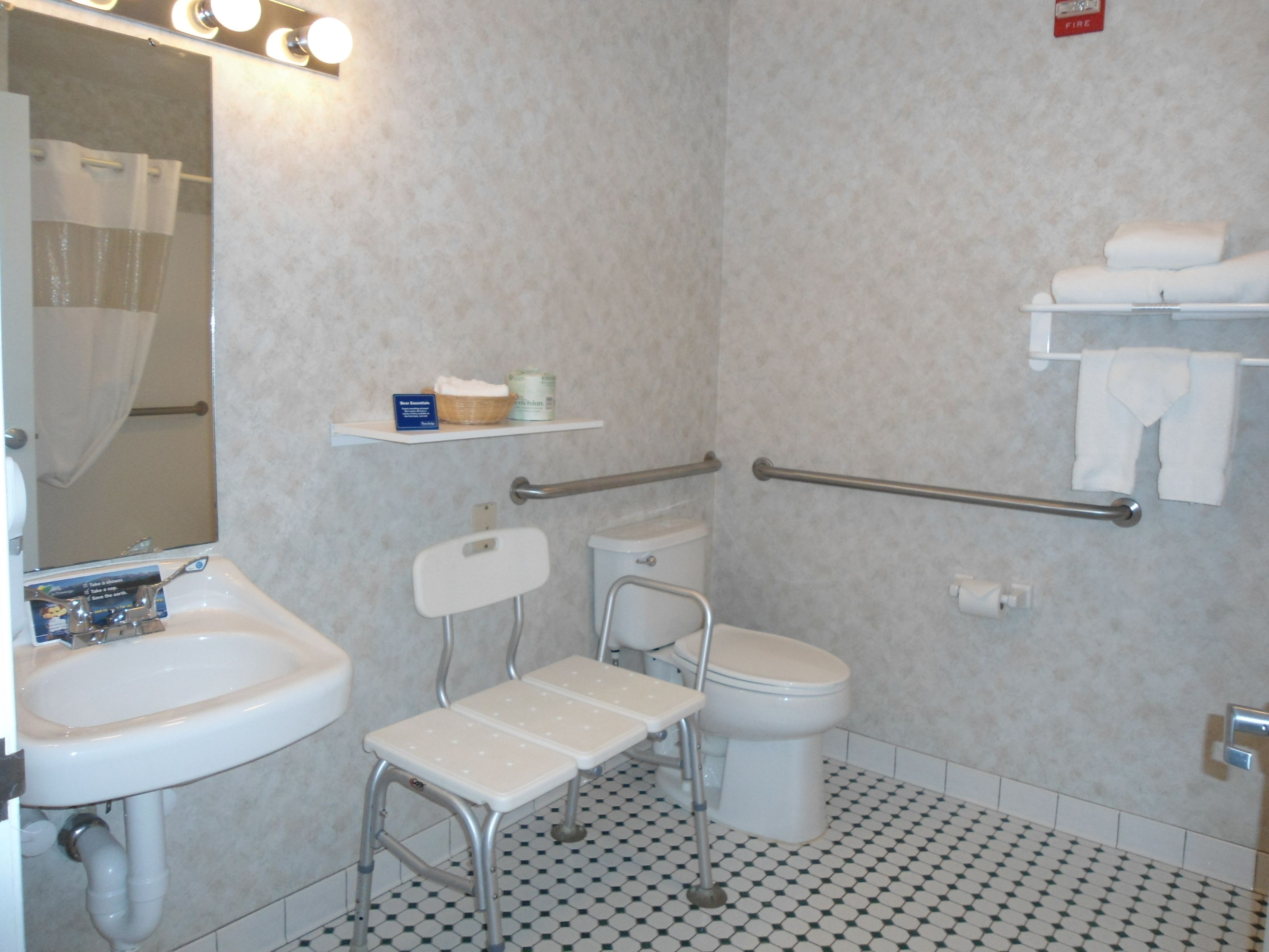 A bathroom with a white sink, a toilet, a shower with a glass door, a white chair, and a shelf with toiletries.