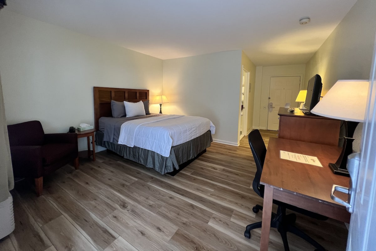 A hotel room with a bed, bedside table, lamp, desk, chair, and a door leading to a bathroom.