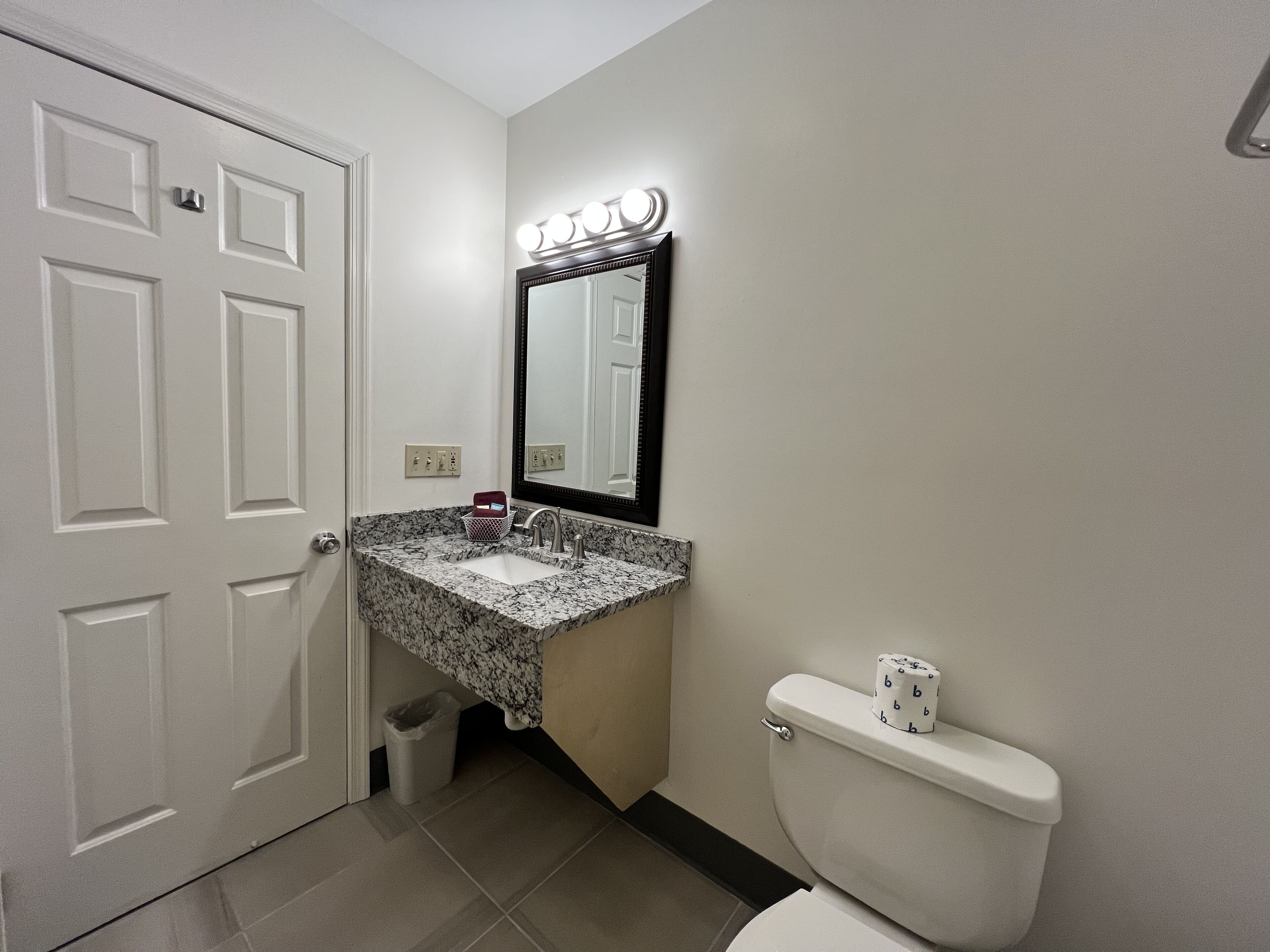A bathroom with a white door, a sink with a marble countertop, a mirror with lights, and a toilet.