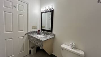A bathroom with a white door, a sink with a marble countertop, a mirror with lights, and a toilet.