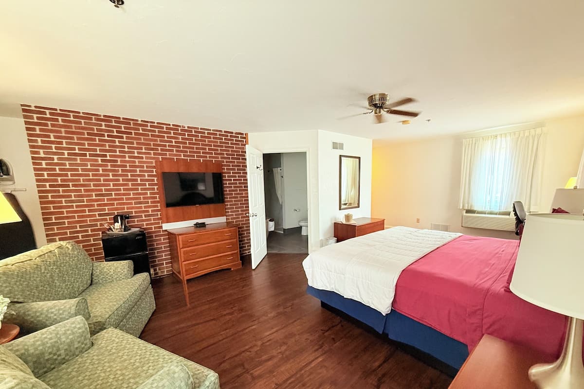 A hotel room with a brick accent wall, a flat-screen TV, a bed with a white and pink bedspread, a wooden dresser, a desk with a lamp, and a ceiling fan.