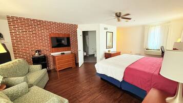 A hotel room with a brick accent wall, a flat-screen TV, a bed with a white and pink bedspread, a wooden dresser, a desk with a lamp, and a ceiling fan.