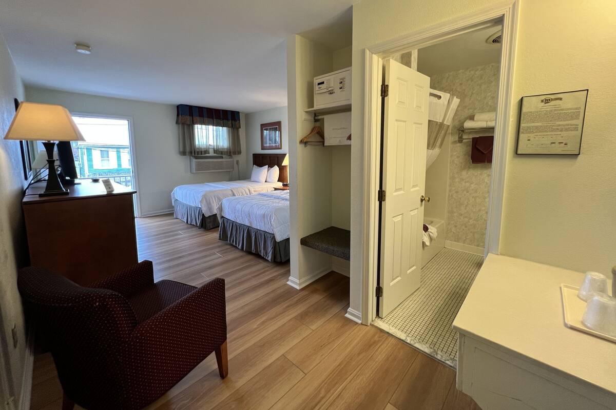A hotel room with two beds, a desk with a lamp, a chair, and a bathroom with a shower and toilet.