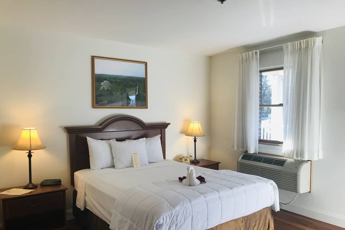 A neatly made bed with a white comforter and pillows, two bedside lamps, a nightstand with a telephone, a framed landscape painting above the bed, and a window with curtains and an air conditioning unit.