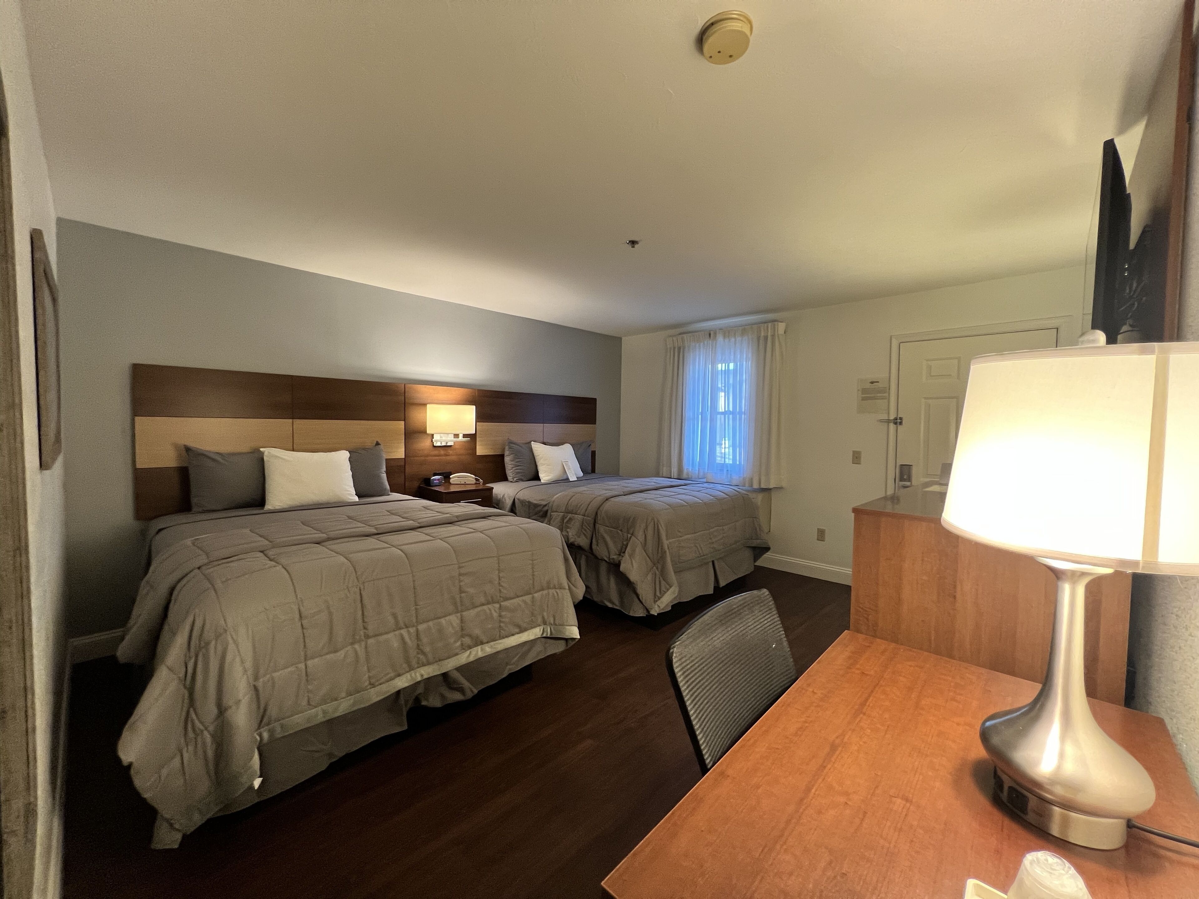 A hotel room with two beds, a desk, a lamp, and a window with curtains.