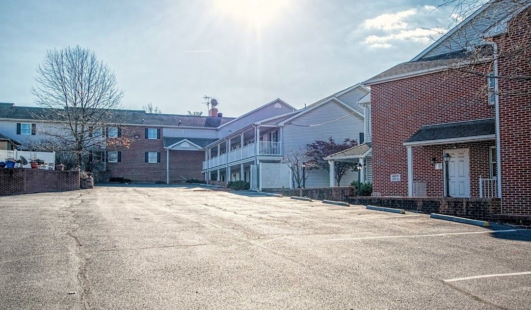 A parking lot with a row of residential buildings, including a two-story house with a balcony and a single-story house with a porch.