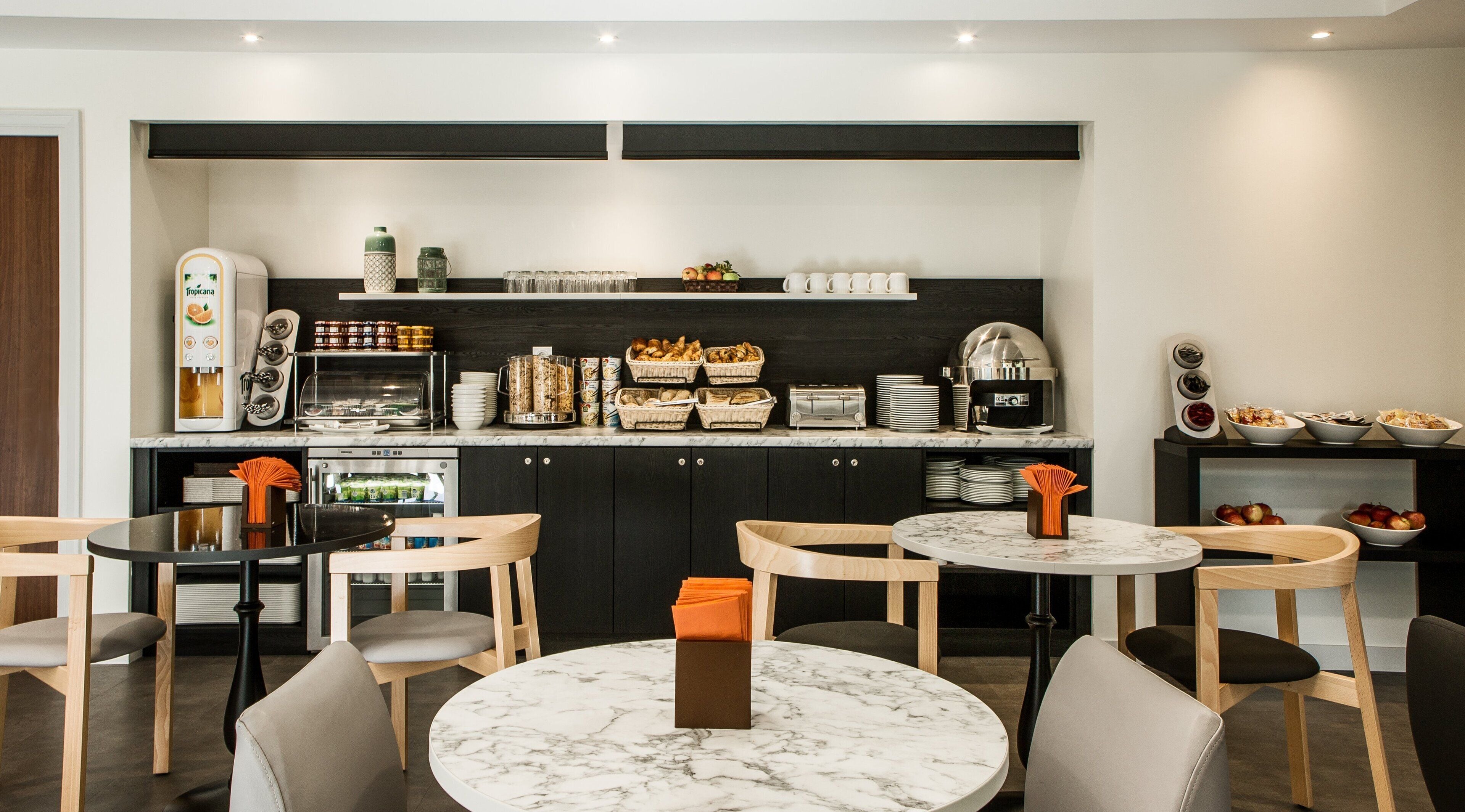A modern kitchen with a built-in coffee station, a toaster, and various food items on shelves. There are four round tables with chairs, and a small refrigerator.