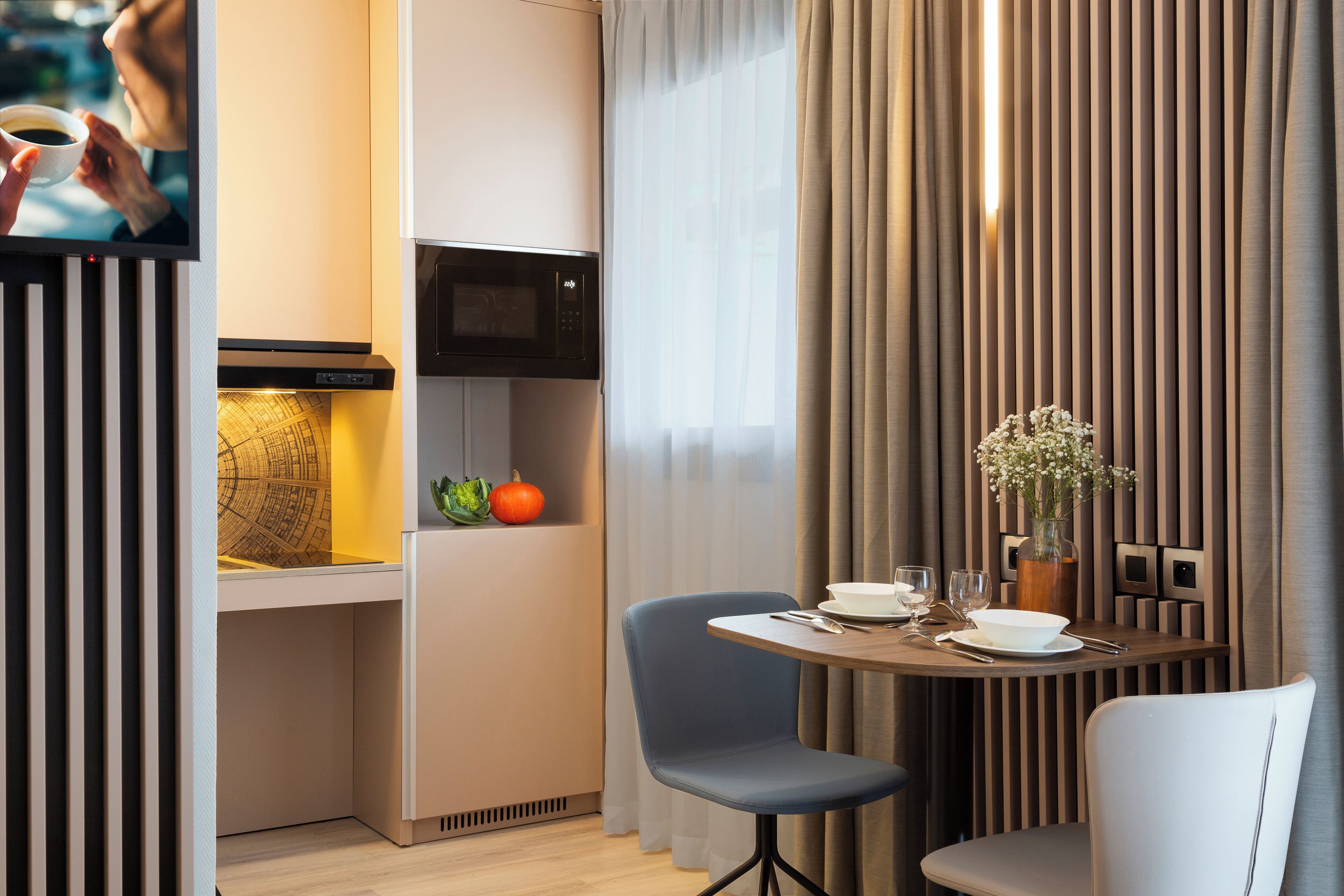 A modern hotel room with a dining area, a microwave, a television, and a small kitchen space.