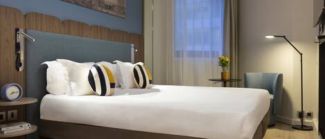 A hotel room with a large bed, a nightstand with a clock and books, a chair, a lamp, and a window with curtains.