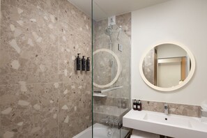 A modern bathroom with a glass-enclosed shower, a round mirror, and a white sink with toiletries. - Residence Inn by Marriott Paris Didot Montparnasse (Paris)
