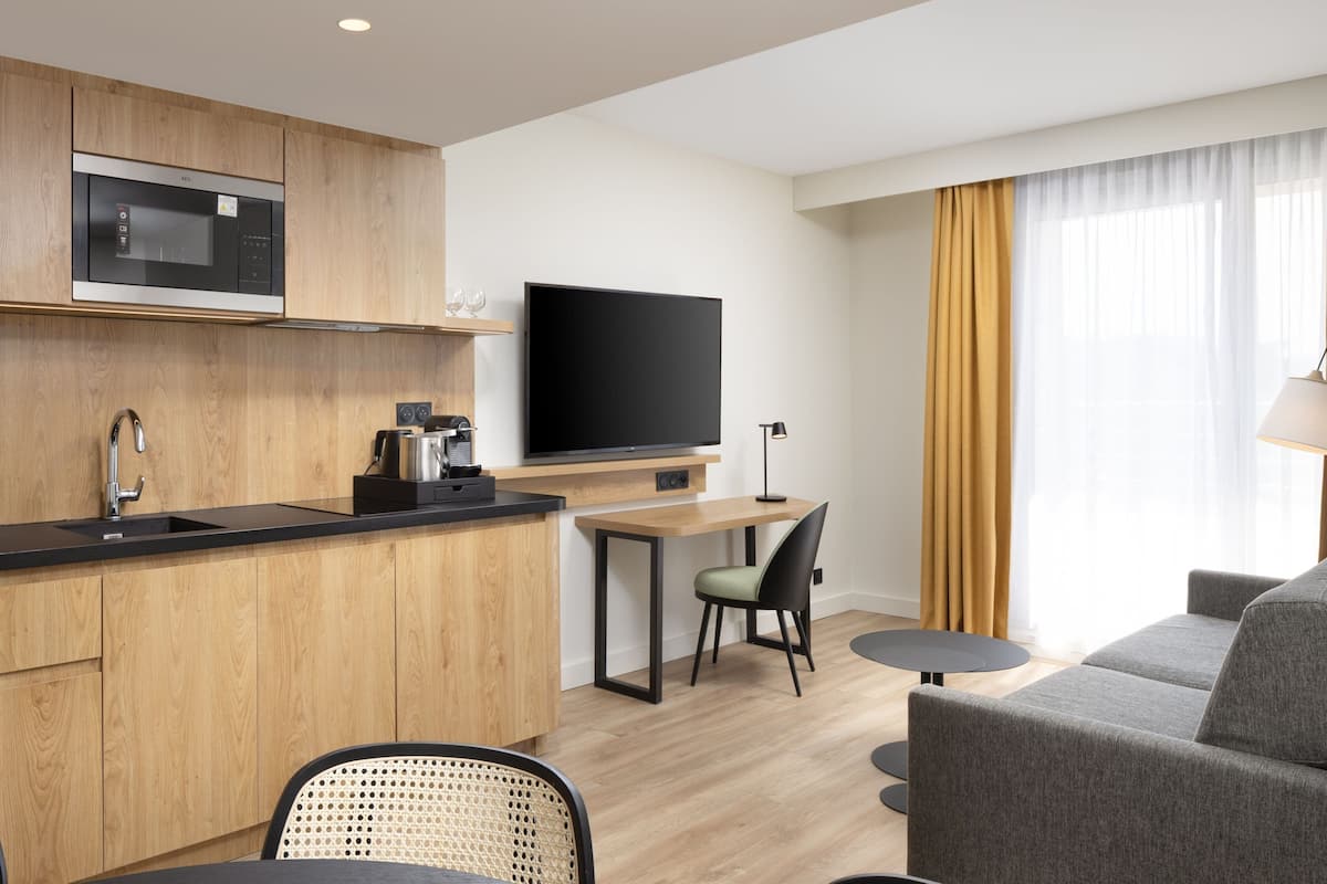 A modern hotel room with a kitchenette, a flat-screen TV, a sofa, and a dining area.