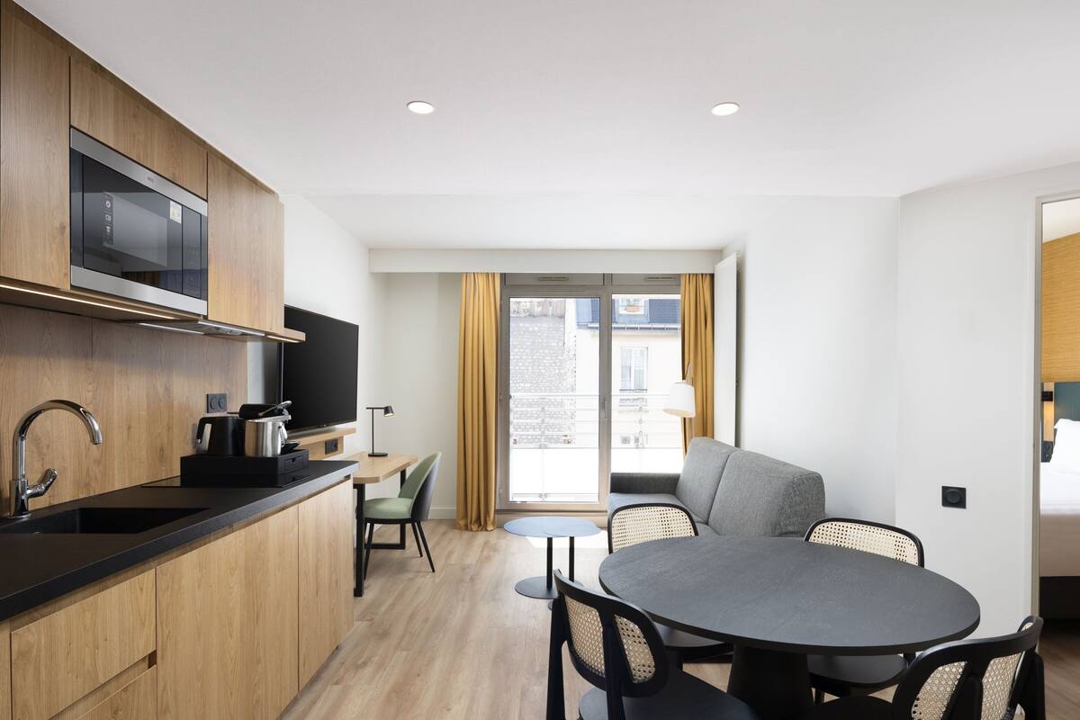 A modern kitchen with wooden cabinets, a built-in oven, and a flat-screen TV. There is a dining area with a round table and chairs, and a large window with curtains.