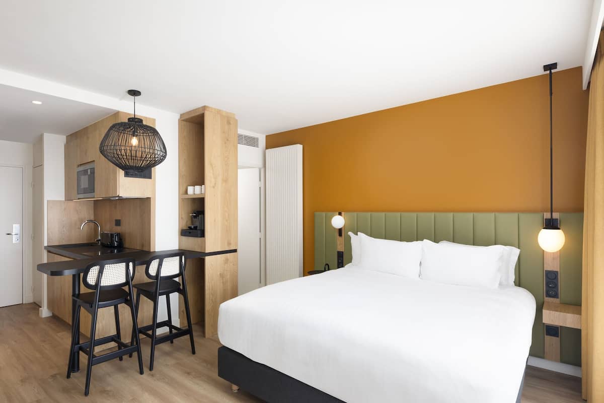 A modern hotel room with a large bed, a kitchenette with bar stools, and a unique green headboard.