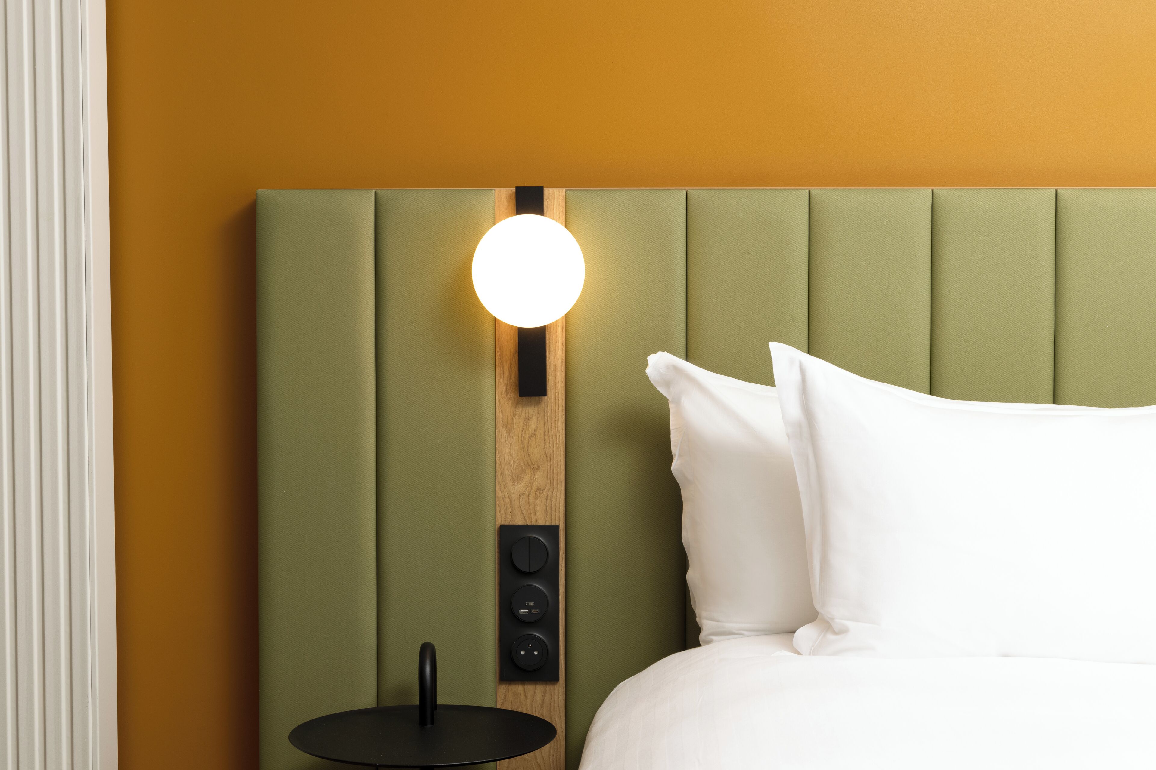 A bed with a green upholstered headboard, a bedside table with a black finish, and a round wall-mounted light fixture.