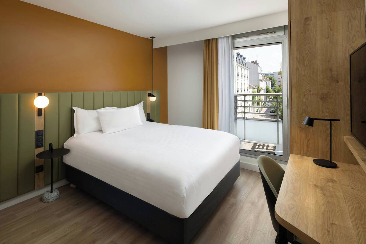 A modern hotel room with a large bed, a wooden desk, and a view of the city through a large window.