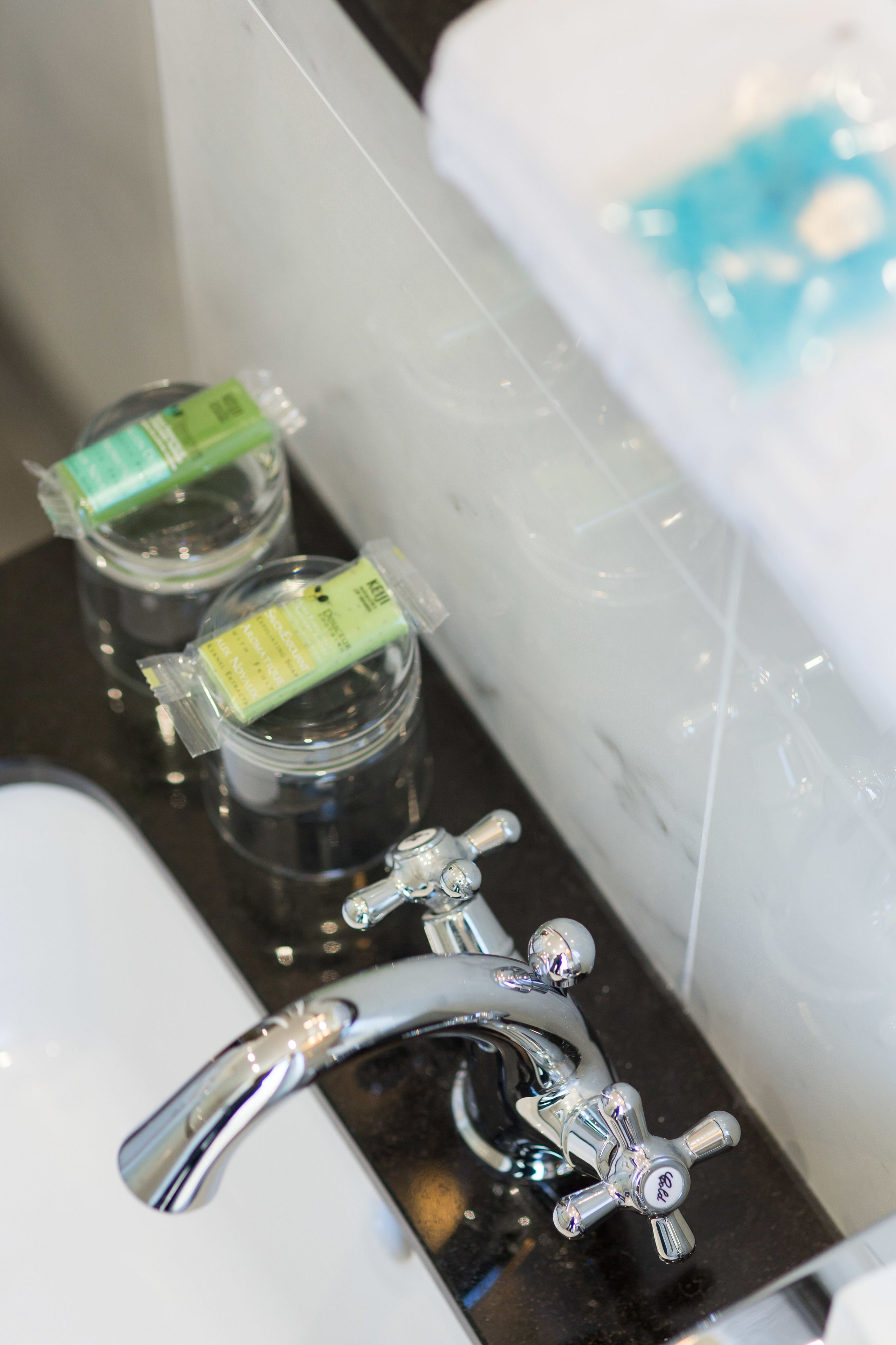 A bathroom with a sink, two glass jars with green labels, a chrome faucet, and a bathtub with a blue and white bath mat.