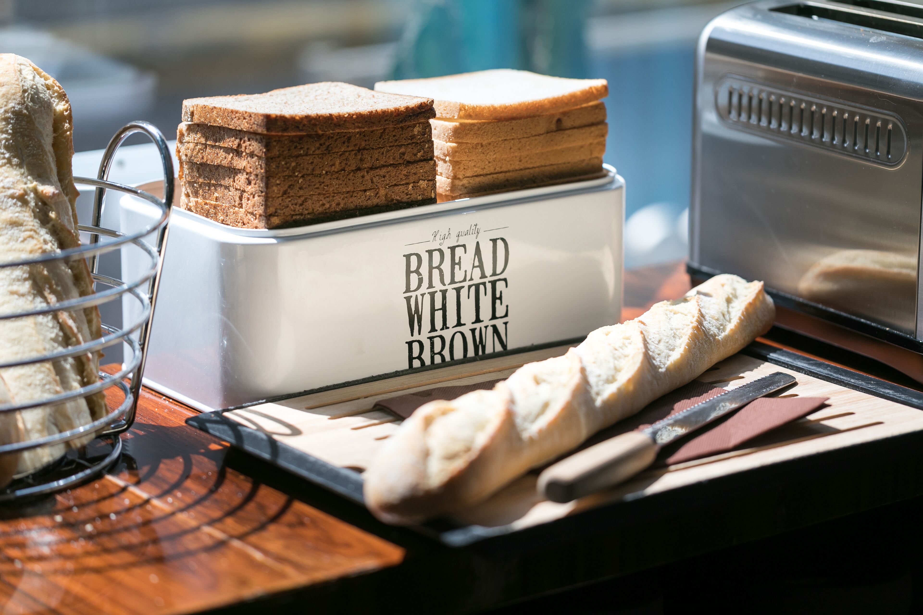 Un grille-pain avec des tranches de pain blanc et de pain complet, un couteau et un panier à pain, le tout posé sur une table en bois.