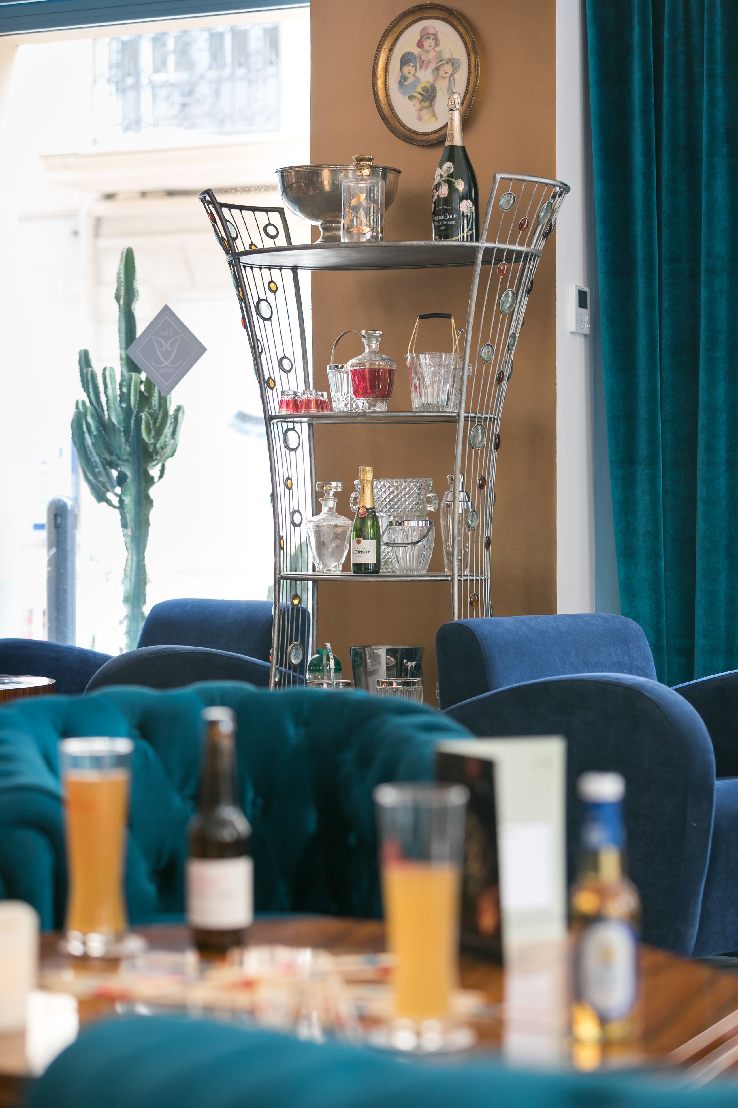 Un comedor con un estante metálico que contiene varias botellas y vasos, una planta de cactus y sillas tapizadas en azul.