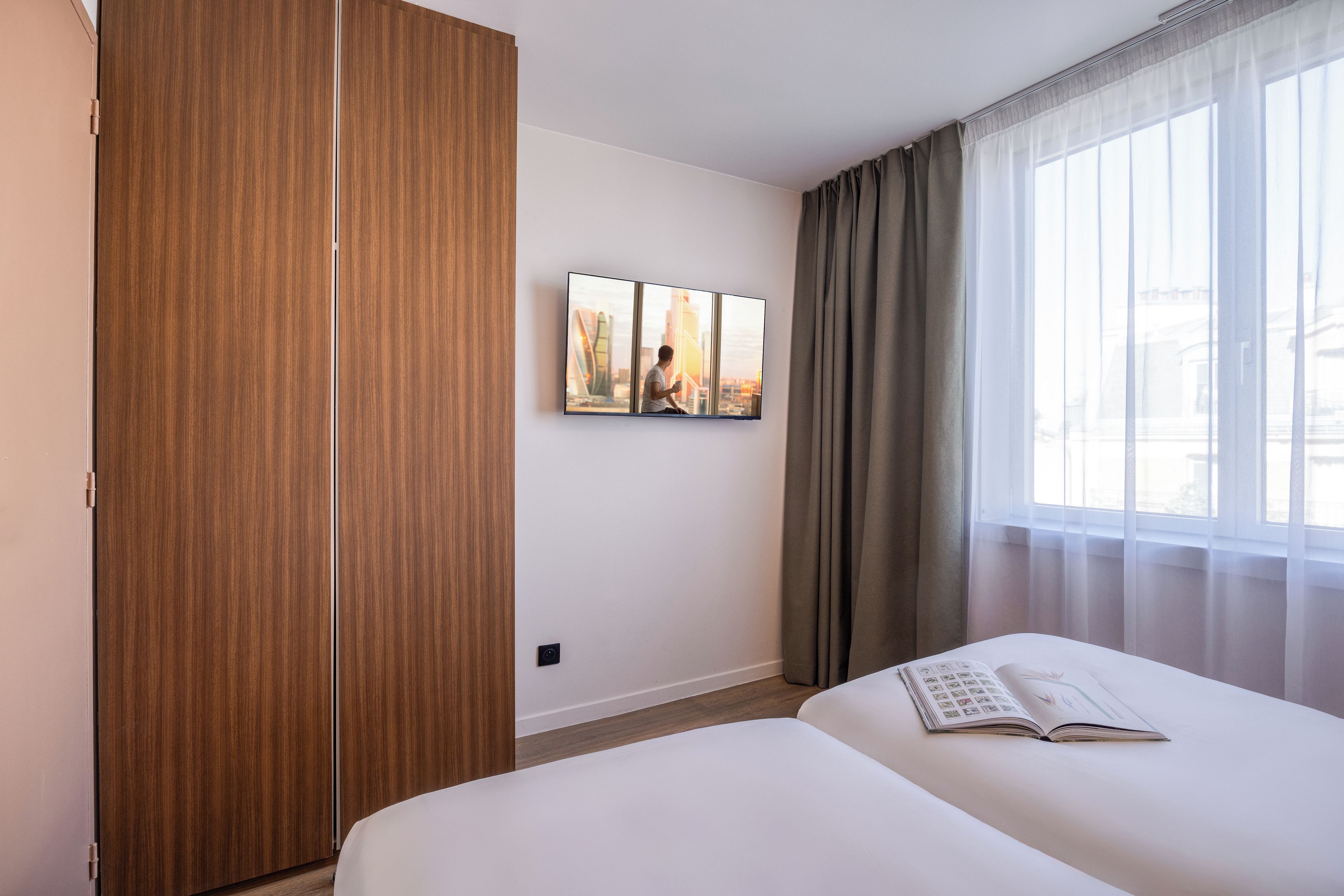 Habitación de hotel con cama, armario, televisor montado en la pared y ventana con cortinas.