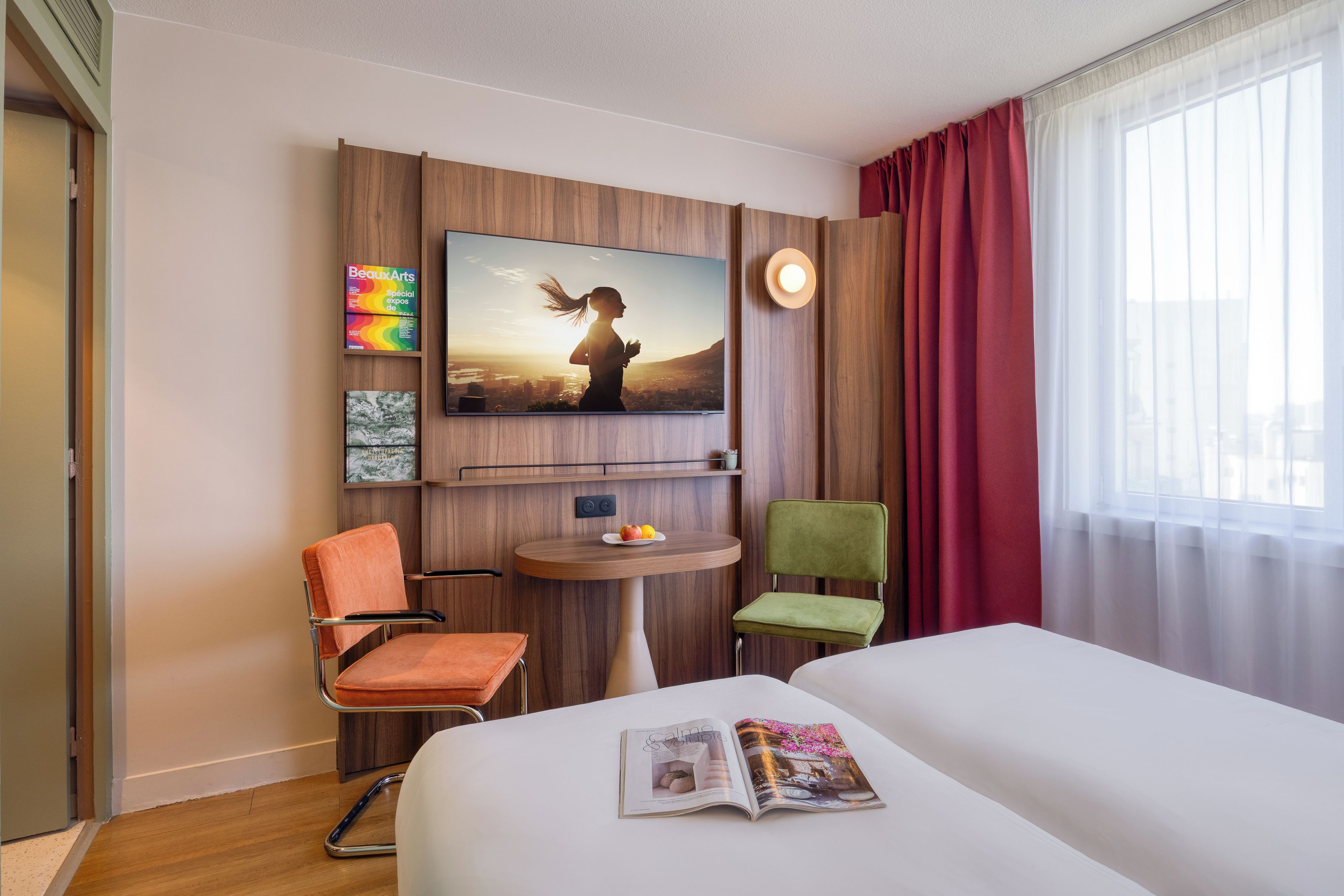 Habitación de hotel con cama, televisor montado en la pared, una mesita, dos sillas y una revista sobre la cama.