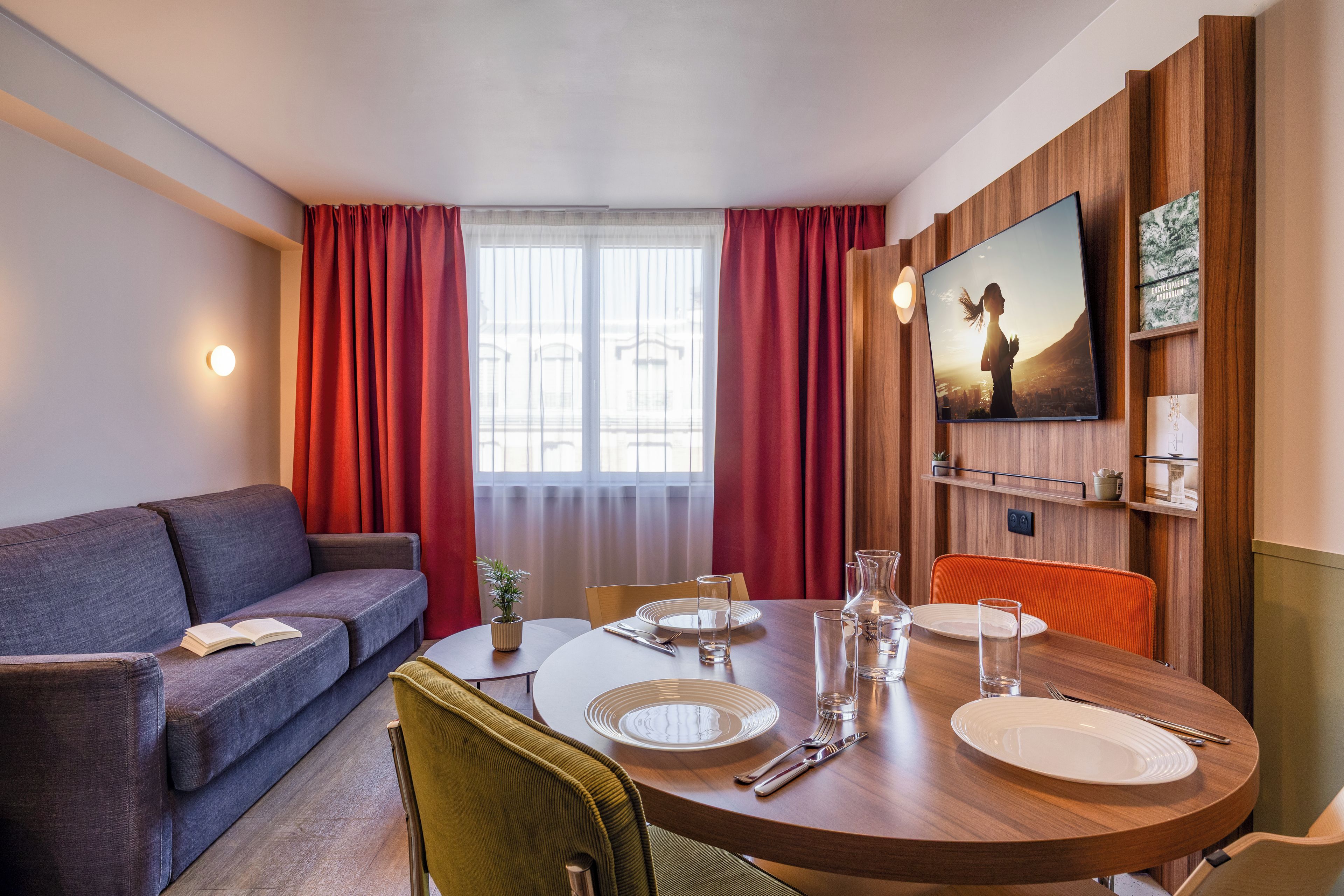 A modern hotel room with a round dining table, a sofa, and a television mounted on the wall.