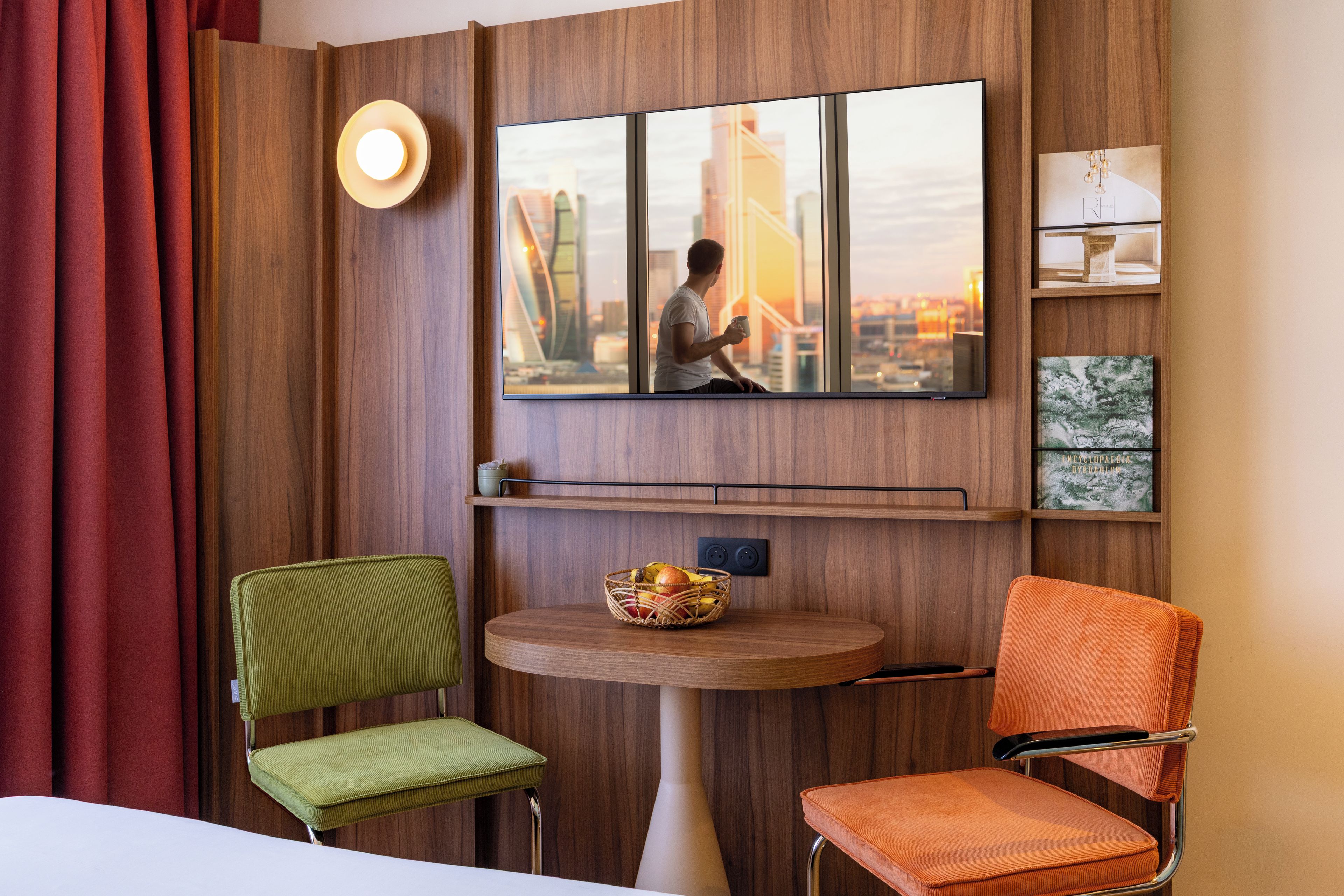 Quarto de hotel com uma janela grande, televisão de tela plana, uma pequena mesa com uma cesta de frutas e duas poltronas estofadas.